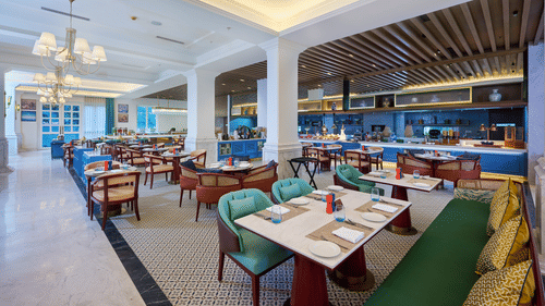 An image of our dining area with fancy tables, cosy chairs, premium couches all placed under chandeliersA far angle shot of cosy tables in a line placed by the side of a buffet setting - Sarovar Premiere, Amritsar