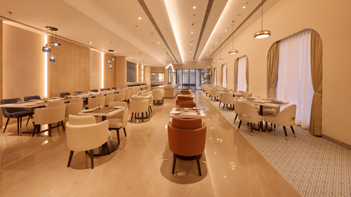 Skywings Sarovar Portico Mathura restaurant corridor with orange and beige chairs, wooden tables, warm recessed lighting, and elegant cream interiors.
