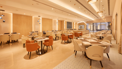 Skywings Sarovar Portico Mathura restaurant with orange and beige cushioned chairs, wooden tables, warm recessed lighting, and cream-toned walls.