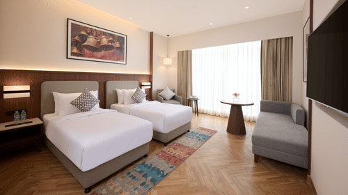 Skywings Sarovar Portico Mathura twin bed room with two single beds, gray sofa, patterned rug, wooden flooring, and warm ambient lighting.