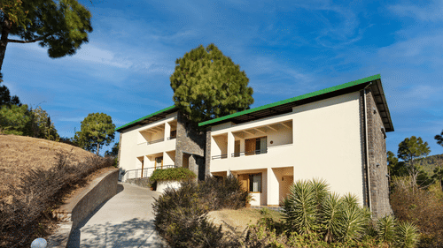 Exterior of premium rooms at Suryavilas Luxury Resort and Spa in Solan featuring the facade of the property on the mountain surrounded by trees and shrubs under a blue sky on a sunny day