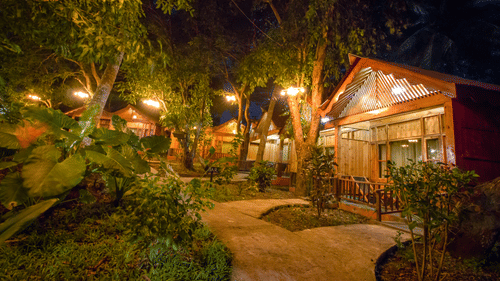 Facade view of a row of wooden cottages during night time with the lights on and greenery next to it - Symphony palms beach resort and spa.