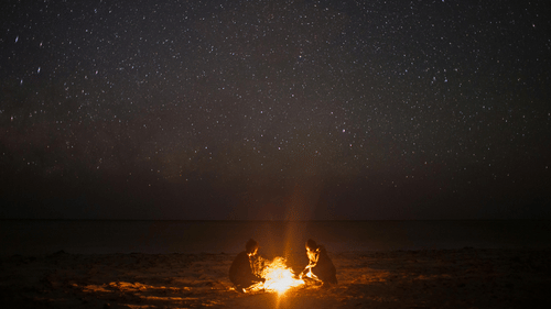 2 people sitting around a bonfire on the beach under a dark sky full of stars.