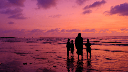 A silhouette of a group of people on a beach at sunset with waves under a purple and pink sky.