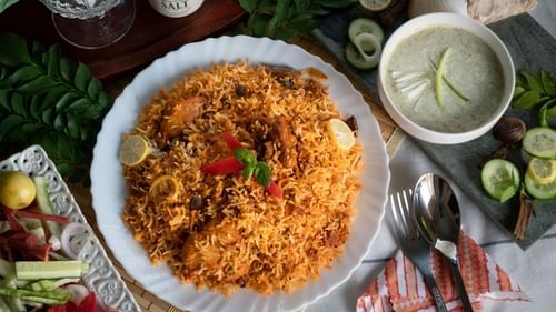 Plate of biryani served with raita, garnished with lime and herbs, surrounded by cut vegetables, spices, and a spoon and fork on a red-striped napkin.
