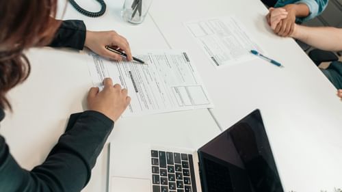 The image shows a person filling out a form at a table. There is a laptop and other paperwork visible on the table as well.