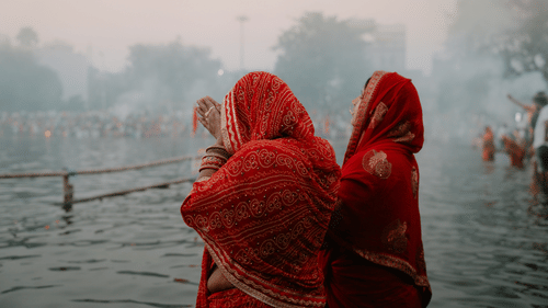 2 women wearing red garments standing in water, with other people visible in the background and a hazy sky above.