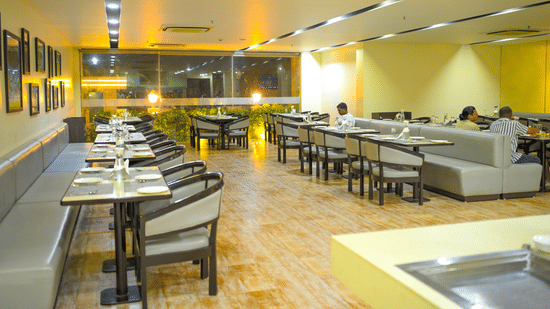 A restaurant dining room at Ambar Sarovar Portico, Gandhidham features neatly arranged tables, chairs, and cushioned booth seating along the wall.