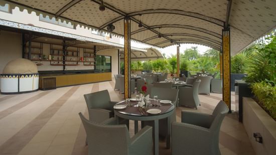 View of the covered outdoor restaurant space showing dining tables, chairs, and a service counter in the background at Arawali Ananta Elite, Jaipur.