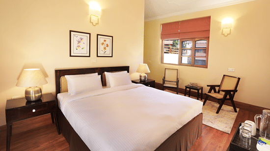 An image of a deluxe room with a double size bed, bedside table with lamps on either side, a chair, and other decorative items - Lamrin Boutique Cottages, Rishikesh