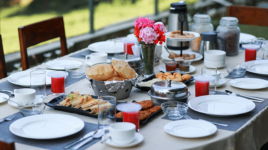 Outdoor breakfast setup with an array of dishes, bread, and beverages on a table surrounded by nature.