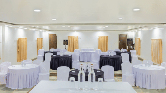 Spacious banquet hall with round tables in white and purple draping, white chair covers, warm recessed lighting and wooden partition walls in background.