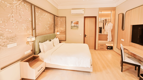 A wide-angle view of a bright guest room at Pride Premier Alwar showing a large bed with white linens, a wall-mounted desk, and a door leading to a private bathroom.