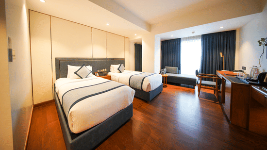 An angled view across the Premium Twin Room at Pride Premier Udaipur. The space displays two single beds arranged in sequence along the wall. A desk with a chair and a wall fixture are positioned on the opposite side near the window area.