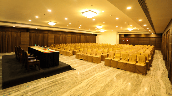 A view of the theatre style seating arrangement at one of the best wedding halls inside Raj Park Hotel, Chennai, featuring chandeliers, a stage and a podium.