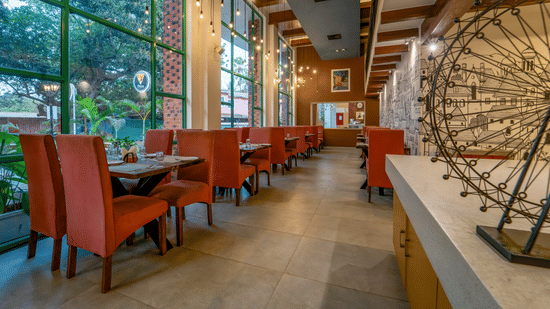Rosetum Anjuna's ground floor restaurant with terracotta high-back chairs, wooden tables, Edison bulb pendant lights, floor-to-ceiling green-framed windows, and a mural wall.