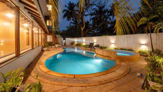 Rosetum Anjuna's kidney-shaped outdoor swimming pool and jacuzzi illuminated at night, surrounded by lush greenery, lounge chairs, and warm wall lighting.
