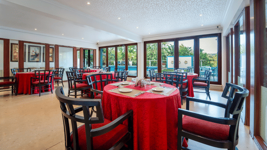 Bright restaurant dining room with red-draped round tables, dark wooden chairs, and large windows overlooking a garden.