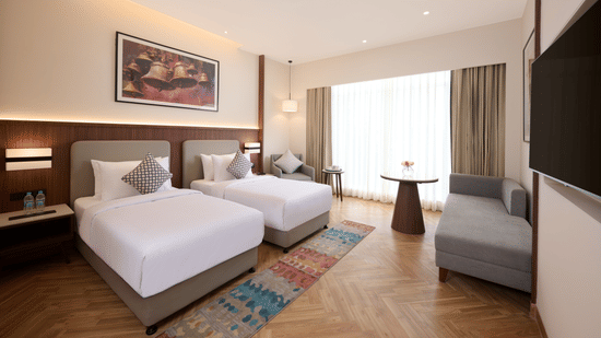Skywings Sarovar Portico Mathura twin bed room with two single beds, gray sofa, patterned rug, wooden flooring, and warm ambient lighting.