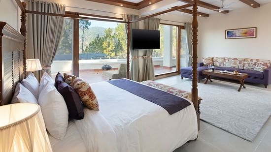 Spacious luxury resort room with a four-poster bed, large windows offering scenic outdoor views, a wall-mounted TV, and a comfortable sitting area with natural light streaming in - Suryavilas Luxury Resort and Spa