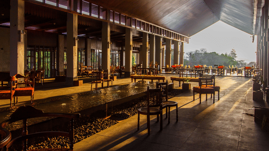 Open-air lounge corridor at Symphony Oceanus Bay Beach Resort & Spa with a long water channel lined with pebbles, wooden chairs, and rows of dining tables lit by the setting sun.
