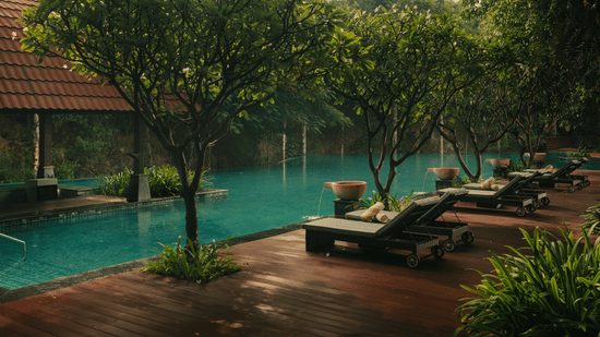 A peaceful view of the swimming pool area at The Ananta Udaipur. The water is a light tone bordered by a deck with several lounge chairs. Lush trees and foliage surround the pool creating a calm atmosphere. A building with a roof is visible in the backgrou