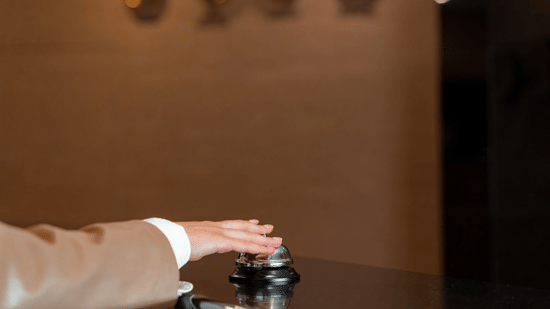 Hotel front desk with two receptionists behind a dark wood counter, ornate red carpet, large framed picture on the wall, and two screens on either side.