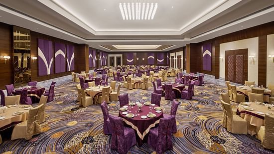 A spacious, elegant banquet hall with round gold-draped tables, purple and gold chairs, and refined place settings. The high ceiling features recessed lighting, a modern chandelier, and purple-gold accents with wood panelling - Voyage The Solitaire Bliss.