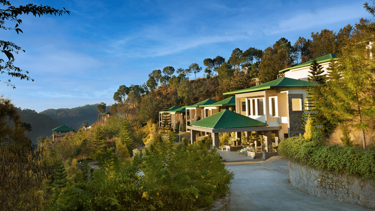 Scenic view of a hillside property surrounded by lush greenery and forested hills under a clear blue sky - Suryavilas Luxury Resort and Spa.