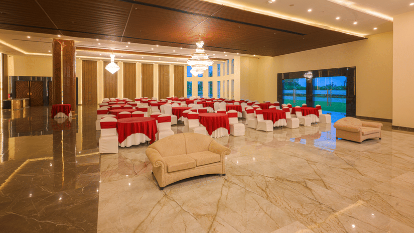 Large banquet hall arranged with round tables, white and red chair covers, decorative lighting and a polished marble floor.