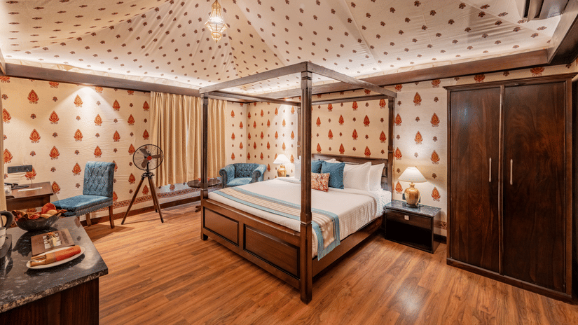 Elegant bedroom with four-poster bed, patterned wallpaper, wooden flooring, wardrobe, blue chairs, and telescope by window.