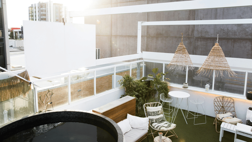 Outdoor urban rooftop terrace featuring a round, dark tub, a seating area with woven furniture, plants, and a partial view of distant buildings.