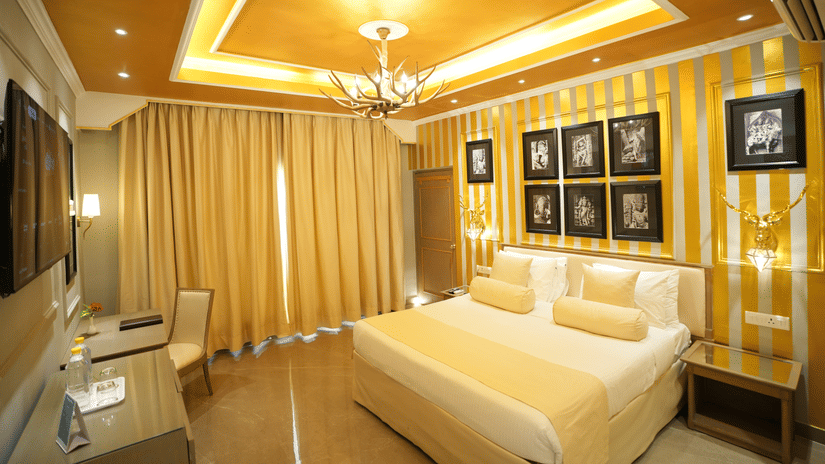 An Executive Suite bedroom at Clarks Khajuraho with a bed, a patterned accent wall with framed artwork, ceiling lighting, and curtains covering a large window.