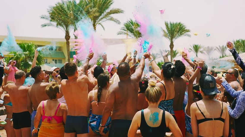 A crowd celebrates at a beach party, tossing vibrant Holi powder into the air.