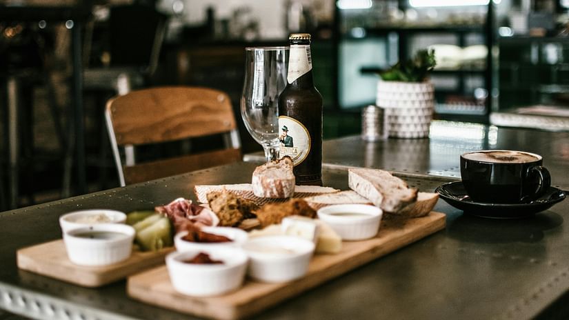 A close-up of a cafe table setting with a wooden platter of assorted snacks, a bottle of beer, an empty glass, and a cup of cappuccino, with a blurred background of a indoor cafe.