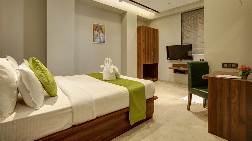 A hotel room featuring a neatly made bed with white linens and green accents, a towel arrangement, wooden wardrobe, desk with chair, wall-mounted TV, and soft ambient lighting - Magnus Fortune House, Magarpatta