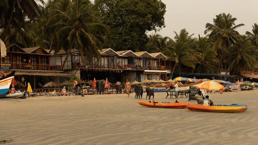 Boats and colorful umbrellas rest on a sandy beach shaded by palm trees and coastal shacks.