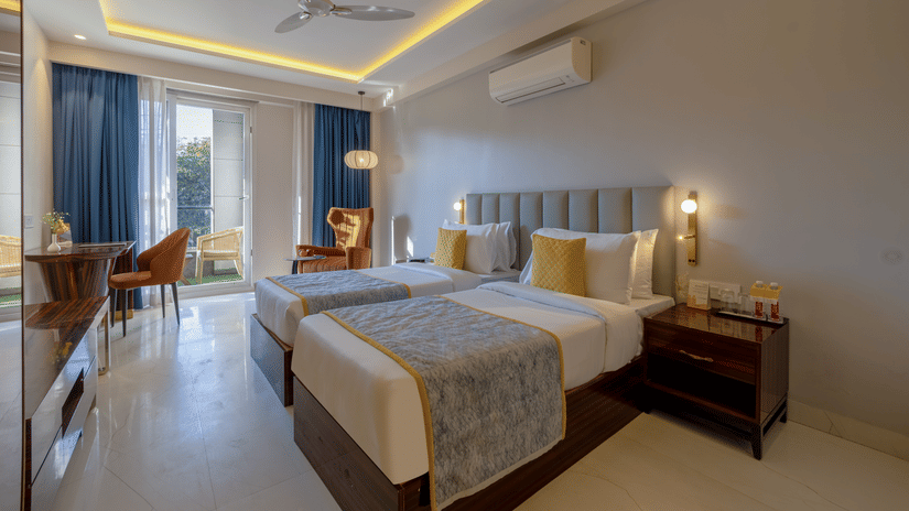Modern guest room at Hotel Bawa, Gurgaon featuring twin beds with yellow accent pillows, blue curtains, a ceiling fan, and warm cove lighting.