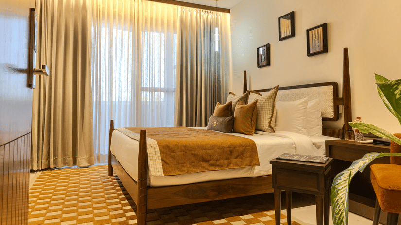 Spacious bedroom with a king size bed dressed in warm toned linen, wooden headboard and soft curtains at Hotel Bawa, Udaipur.