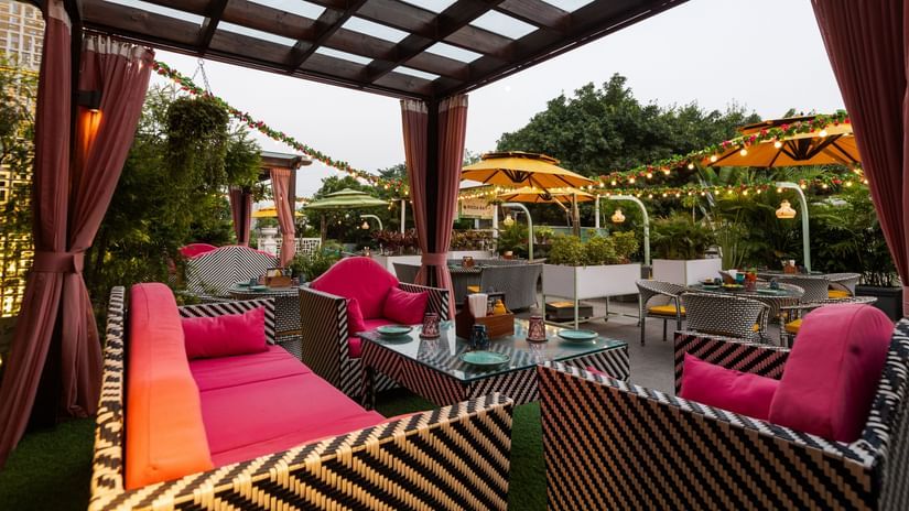 A patio restaurant with outdoor seating featuring a zigzag pattern. A wooden pergola covers the area, which is lit by string lights and includes umbrellas and plants, creating a pleasant dining atmosphere - Karma Lakelands.