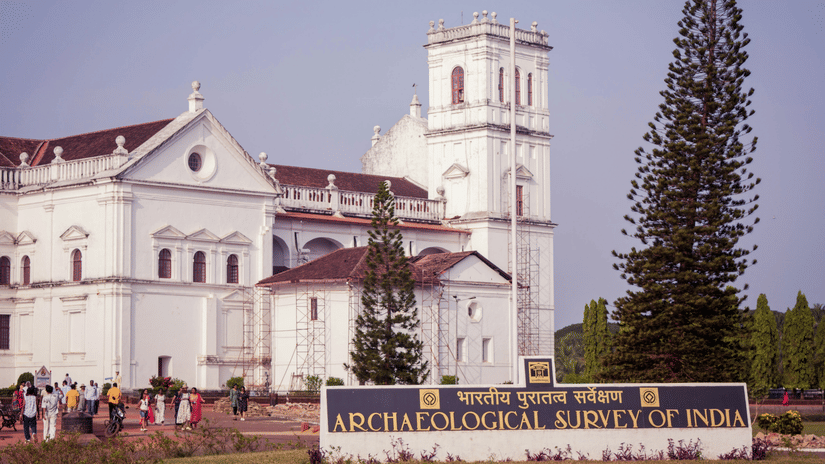 A facade view of Se Cathedral Church in old Goa with many people walking on one side and a large Christmas tree on the other side, there is also a signage with the words Archaeological Survey of India in the foreground.