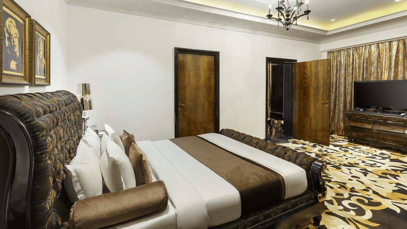 The spacious presidential suite bedroom with a king size bed, upholstered headboard, bedside tables, chandelier lighting, television unit and patterned flooring at La Marvella, Bengaluru.