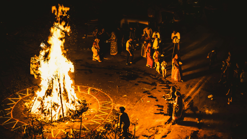 People gathered around a large bonfire at night, standing in a circle as flames rise upward.