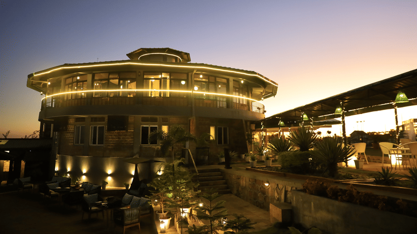 Polo Resort Cherrapunji exterior at twilight with illuminated round main building, outdoor lounge seating, potted plants and pergola covered terrace.