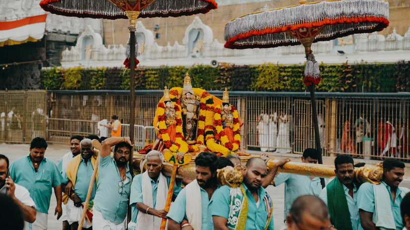 Men carrying a decorated Hindu deity in a colourful ceremonial procession under large umbrellas.