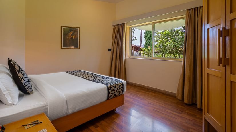 A room with wall hangings, and a wooden almirah and a spacious bed facing a large window with lush greenery – Ras Resort by Treat