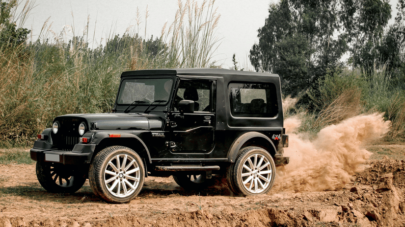 A black Mahindra Thar SUV drives on a dirt road, kicking up a cloud of dust behind it. 