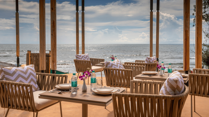 A well arranged tables and chairs at In-House Restaurant with sea waves crashing on the beach in the background