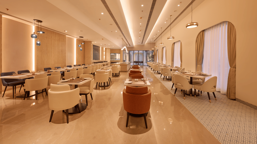 Skywings Sarovar Portico Mathura restaurant corridor with orange and beige chairs, wooden tables, warm recessed lighting, and elegant cream interiors.