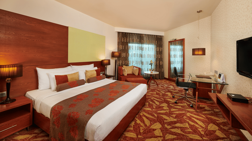 Room at Pride Premier Nagpur featuring a plush bed with pillows and cushions placed in front of a TV and a sitting space next to the window